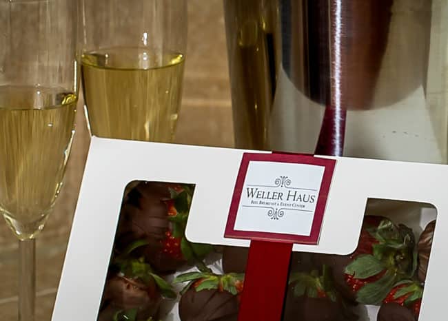 Two glasses of champagne with a white box tied with a red ribbon containing chocolate covered strawberries.